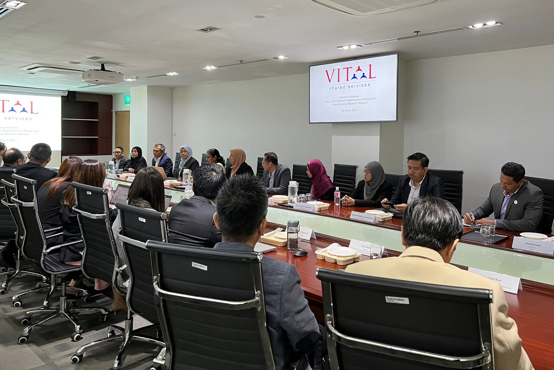 A group of professionals seated around a large conference table with a VITAL Shared Services presentation on two screens.